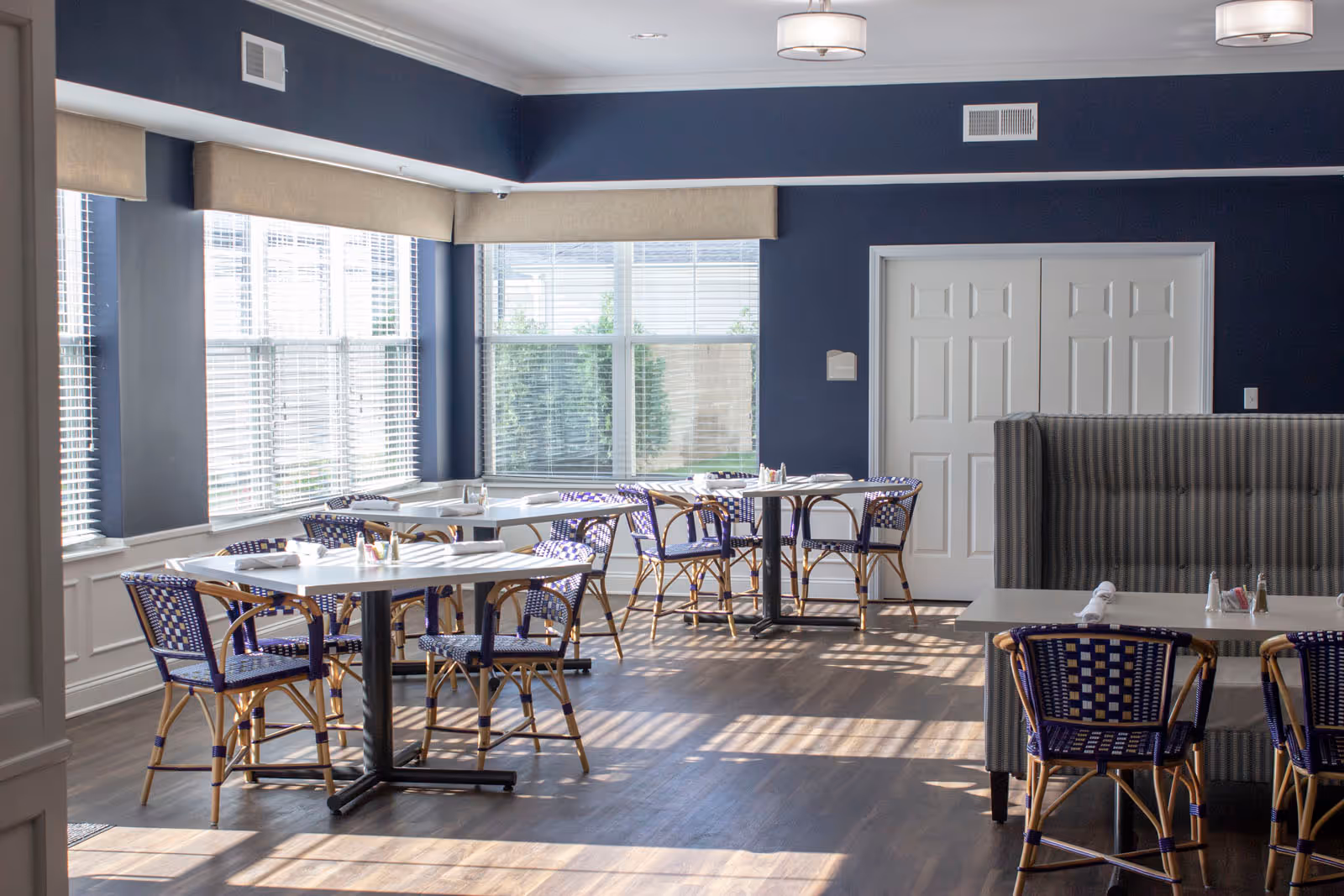 Bright dining room with multiple tables and chairs featuring blue and natural wood woven seats. Large windows with blinds allow natural light to fill the room. The walls are painted dark blue with white trim, and there is a gray upholstered booth seating area on the right side.