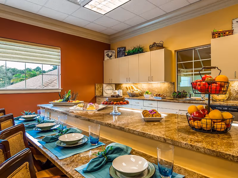 A bright and inviting kitchen and dining area with a marble countertop island set with plates, bowls, glasses, and teal napkins. The kitchen has white cabinets, a window with a view of trees and rooftops, and various food items including fruit baskets and baked goods displayed on the counter.