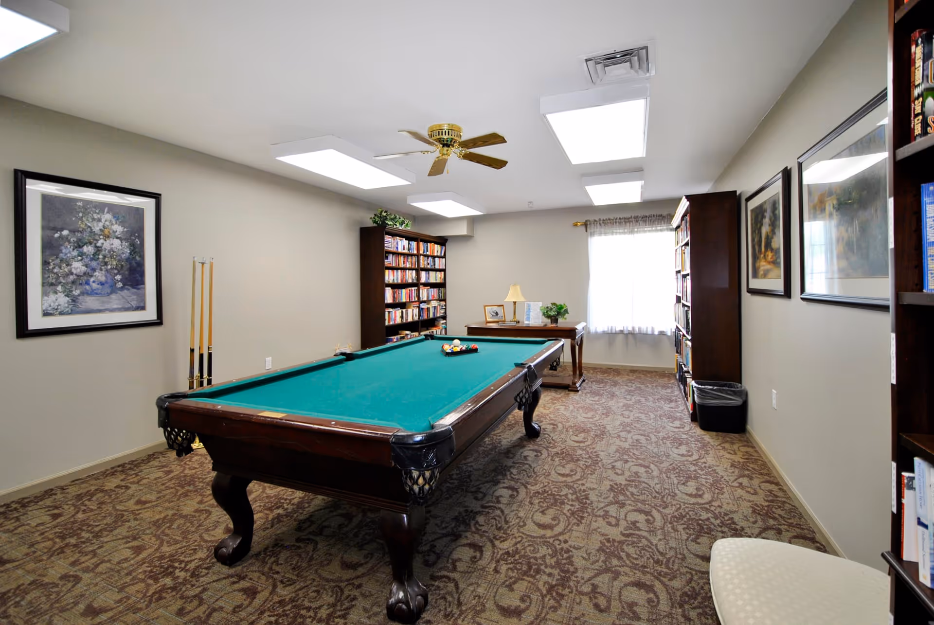 A well-lit room with a green felt pool table in the center, surrounded by bookshelves filled with books. The room has beige walls, framed artwork, a ceiling fan, and a window with sheer curtains letting in natural light. There is a small table with a lamp and decorative items near the window.