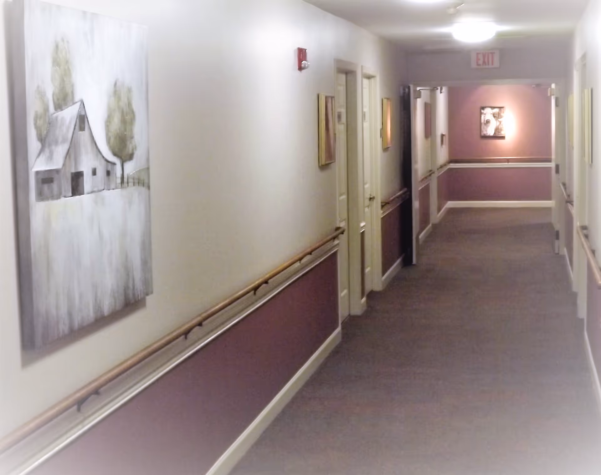 Carpeted interior hallway with handrails, doors and framed artwork leading to an exit sign