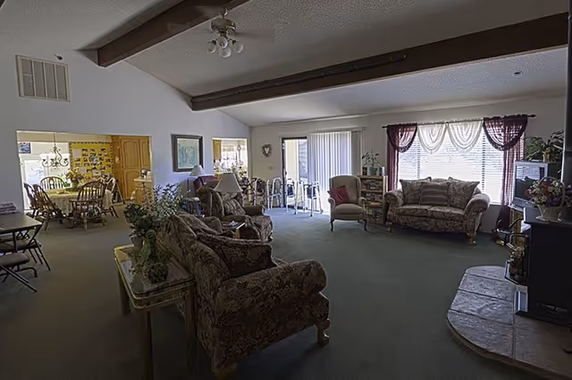Large furnished common living area with patterned sofas and armchairs, coffee tables, a fireplace, and a dining area visible in the background.