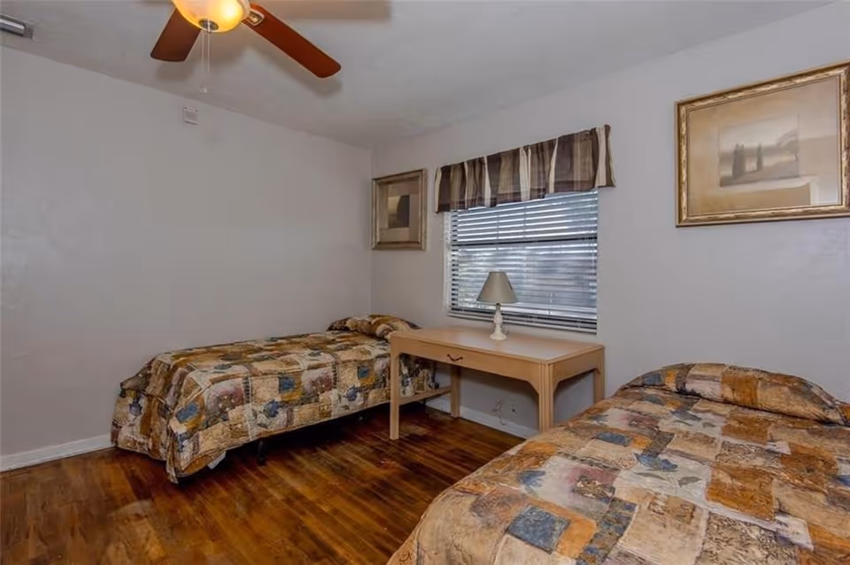 A bedroom with two twin beds covered in patchwork quilts, a wooden floor, a ceiling fan with light, a window with blinds and a valance, a wooden desk with a small lamp, and two framed pictures on the walls.
