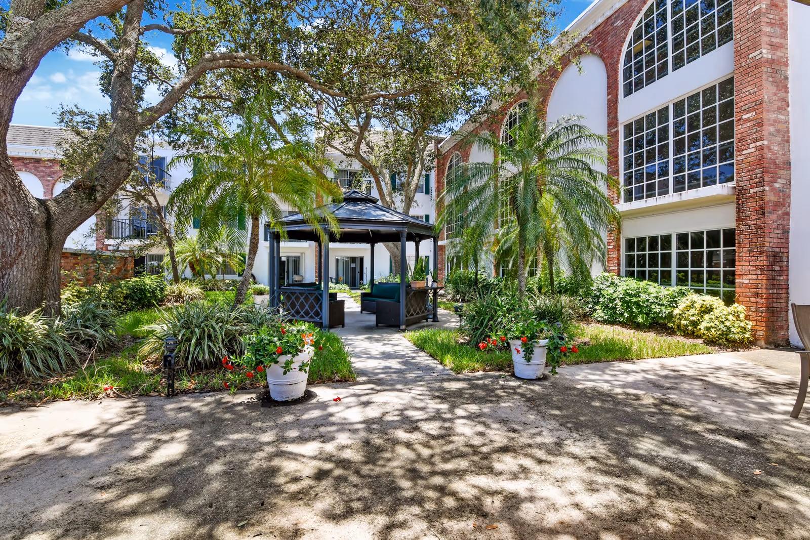 Courtyard with a central gazebo surrounded by palm trees, potted flowers, and a brick-and-white multi-story building.