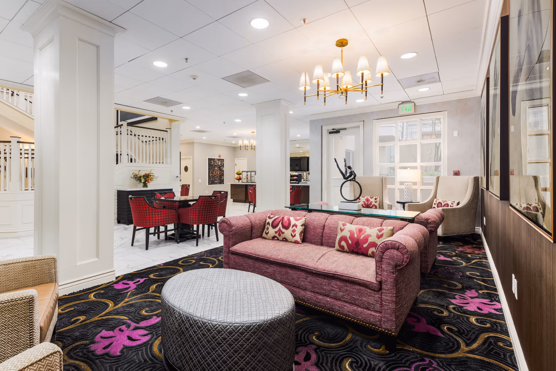 A bright and elegant senior living facility common area featuring a plush purple sofa with patterned pillows, a round gray ottoman, two beige armchairs with patterned cushions, and a glass coffee table with a modern sculpture. The room has white pillars, a black carpet with purple and gold floral designs, and a chandelier with multiple lampshades. In the background, there is a dining area with red cushioned chairs and tables, and a staircase with white railings.