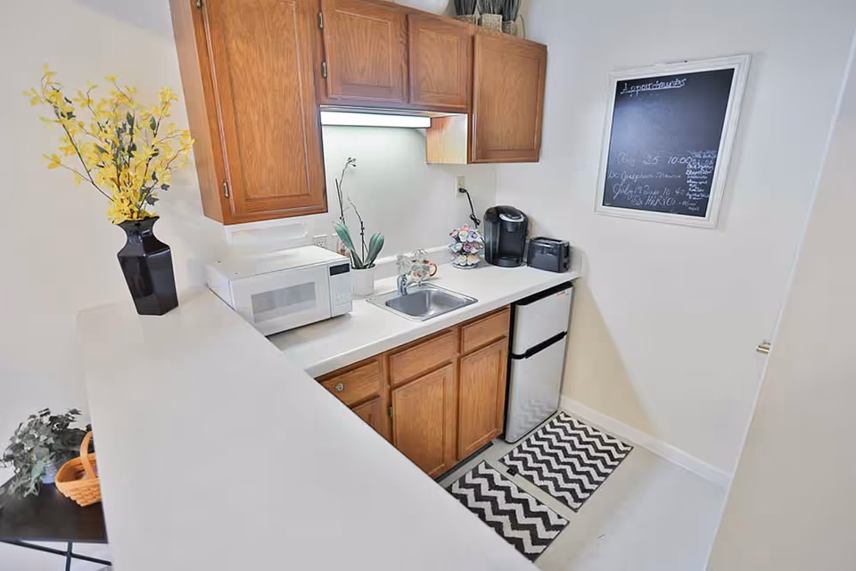 Small kitchen area with wooden cabinets, a white countertop, a microwave, a small sink, a coffee maker, a toaster, a mini refrigerator, and a black vase with yellow flowers. A blackboard with handwritten notes is mounted on the wall.