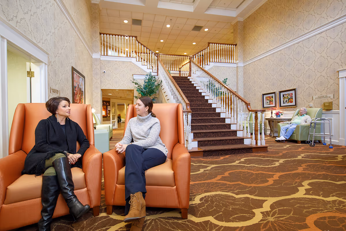Two women sitting and talking in orange armchairs in a spacious, well-lit senior living facility lobby with patterned wallpaper and carpet. A large staircase with white railings and wooden handrails is in the background. An elderly woman sits in a green armchair near a walker on the right side of the image.