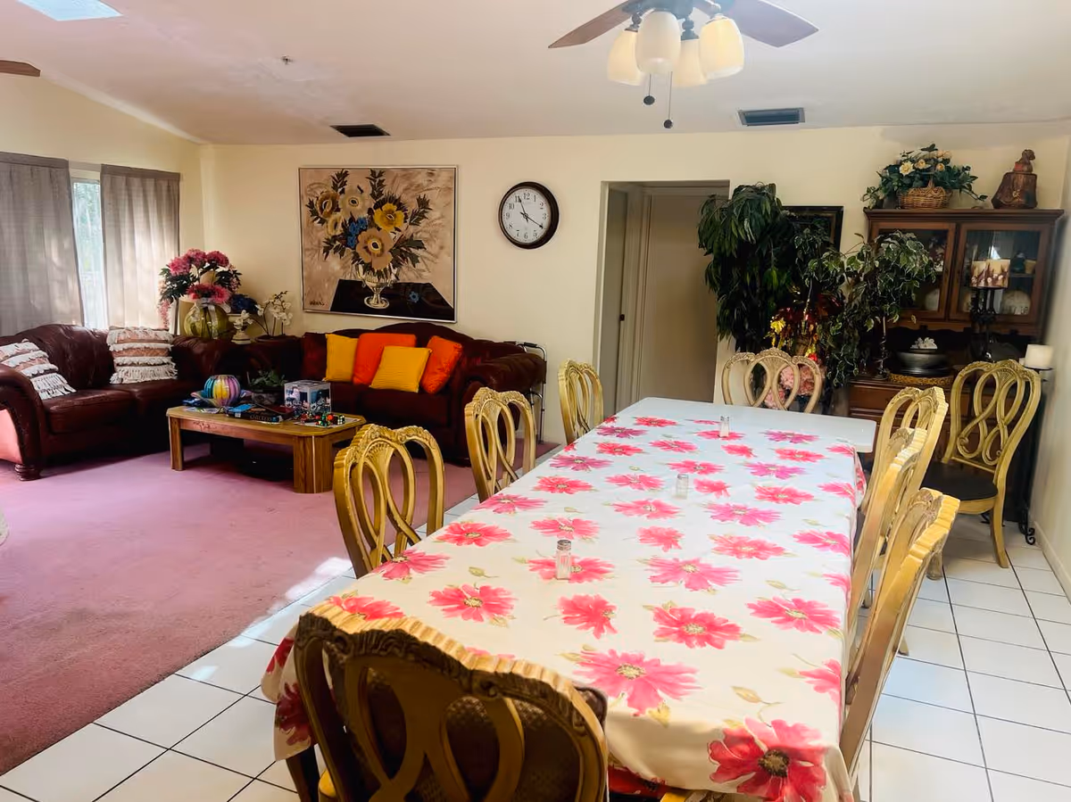 A combined living and dining area in a senior living facility. The dining table is covered with a white tablecloth featuring large pink flowers and surrounded by ornate wooden chairs. The living area has a brown leather sectional sofa with colorful cushions and a wooden coffee table. The room is decorated with plants, a floral painting, a clock on the wall, and a wooden cabinet with decorative items. The floor is tiled near the dining area and carpeted in the living area.
