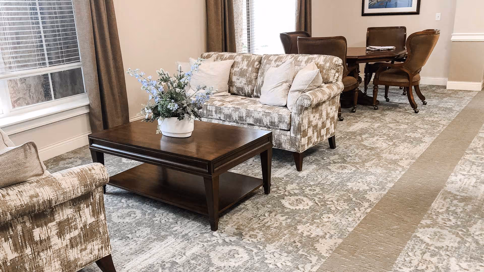A cozy living room area with patterned beige sofas, a dark wooden coffee table with a white vase of blue and white flowers, and a dining table with four brown leather chairs in the background. The room has large windows with brown curtains and a patterned carpet in neutral tones.