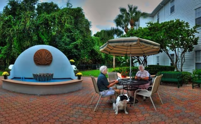 Two elderly people sit at a patio table under an umbrella with a dog nearby beside a decorative fountain in a courtyard outside a residential building.
