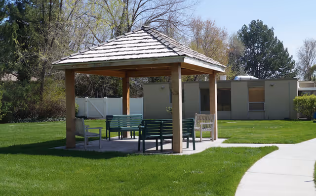 A wooden gazebo with benches on a green lawn beside a sidewalk and a low single-story building.