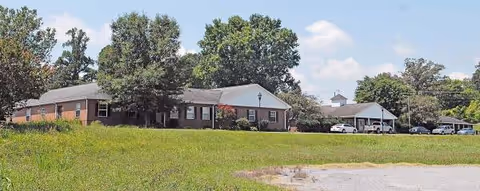 Single-story brick senior living campus with green lawn, trees, and parked cars.