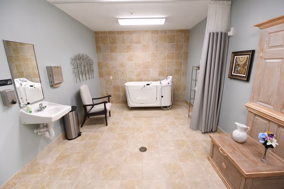 A spacious bathroom with beige tiled floor and wall, featuring a white walk-in bathtub against the back wall. On the left side, there is a wall-mounted sink with a mirror above it, a soap dispenser, a paper towel dispenser, a trash can, and a cushioned armchair. On the right side, there is a wooden cabinet with a white pitcher and a small vase of flowers on top, a framed picture on the wall, and a gray privacy curtain.