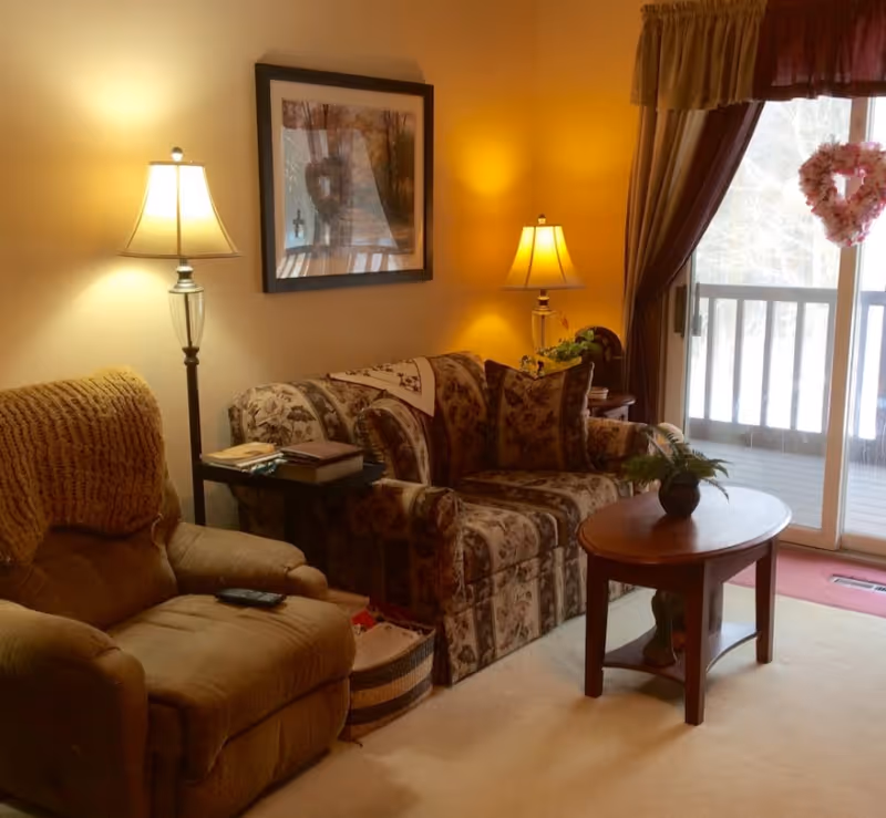 Cozy living room with a floral patterned sofa, a brown recliner chair with a knitted throw, two lit table lamps, a wooden coffee table with a small potted plant, a side table with books, a framed picture on the wall, and a sliding glass door with curtains leading to an outdoor balcony.