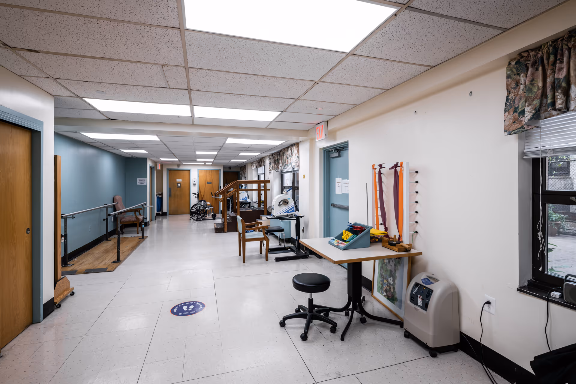 A hallway in a nursing and rehabilitation center with handrails along the walls, a wheelchair, exercise equipment, chairs, and a table with therapy tools. There are windows with floral curtains on the right side and doors at the end of the hallway.