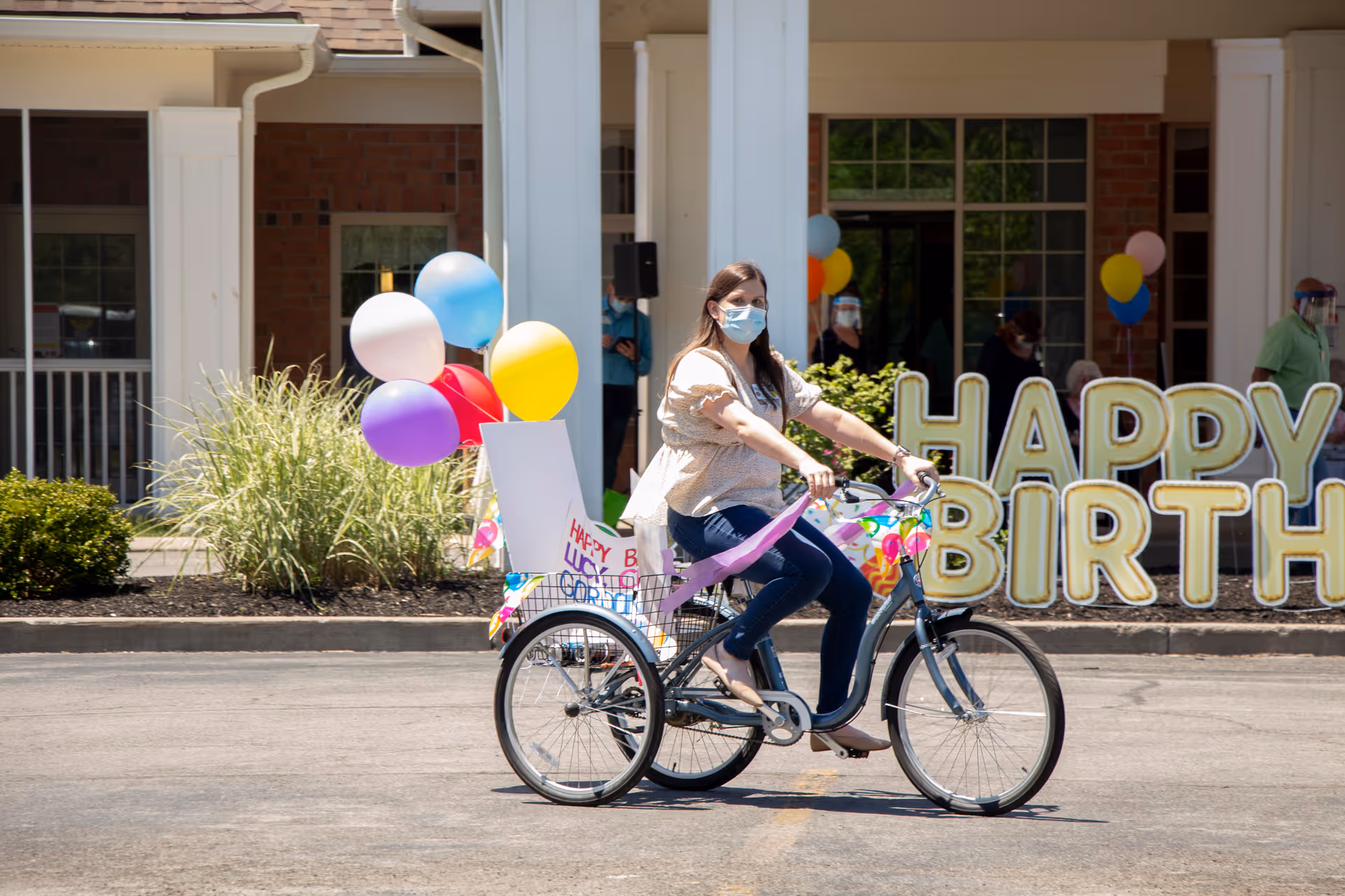 A woman wearing a face mask rides a decorated tricycle with colorful balloons and a birthday sign in front of a building entrance. Large gold balloons spelling out 'HAPPY BIRTHDAY' are displayed in the background, with a few people standing nearby, some wearing face shields.