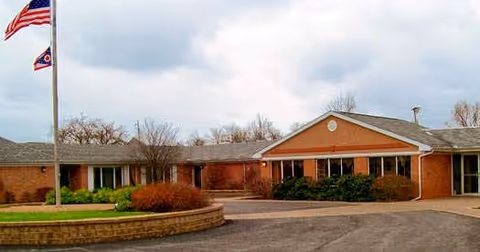 Exterior view of a single-story brick building with a gabled roof, surrounded by shrubs and a circular brick planter in front. Two flags, including an American flag, are flying on flagpoles to the left. The sky is cloudy.