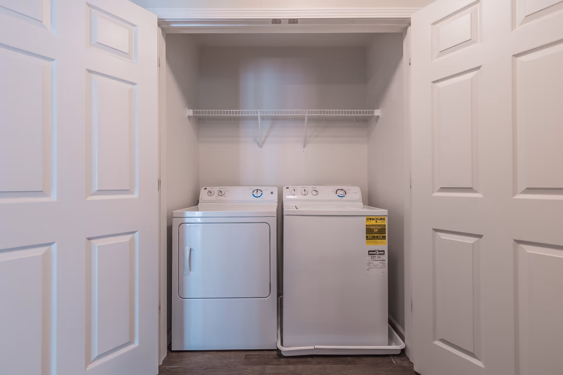 A laundry area with a white washing machine and dryer side by side inside a closet with double white doors. There is a white wire shelf above the machines and the walls are painted light gray.