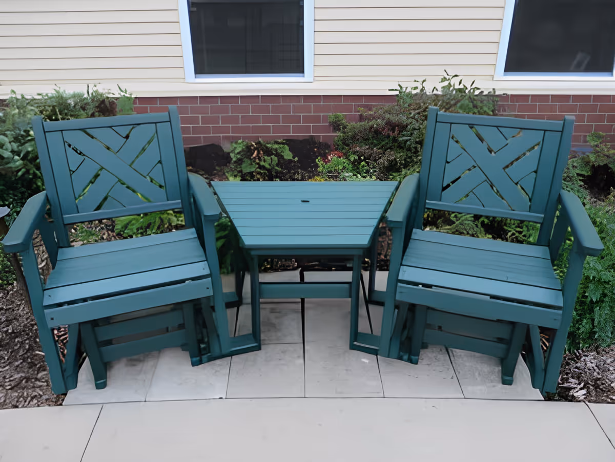 Two teal outdoor chairs with armrests and a matching small table between them on a tiled patio in front of a building with beige siding and two windows, surrounded by green shrubs and plants.