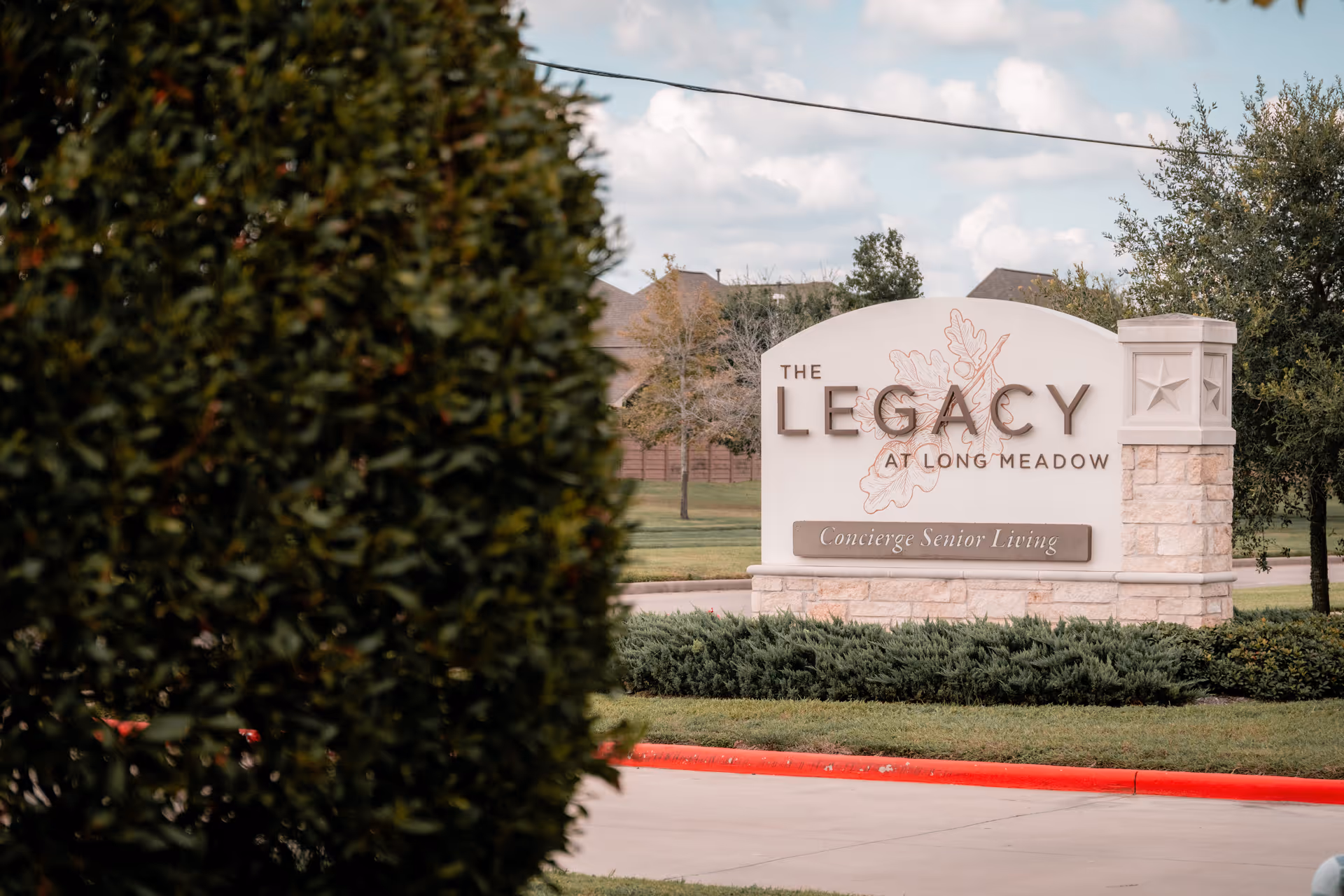 Outdoor view of a large stone sign for The Legacy at Long Meadow, a concierge senior living facility, surrounded by greenery and trees with a partly cloudy sky in the background.