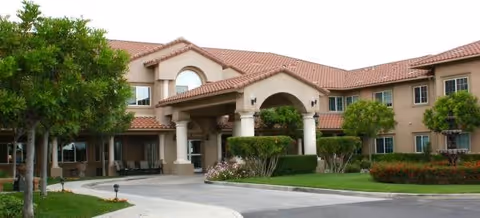 Exterior view of a two-story senior living facility building with a tiled roof, arched entrance supported by columns, surrounded by green trees, shrubs, and a well-maintained lawn.