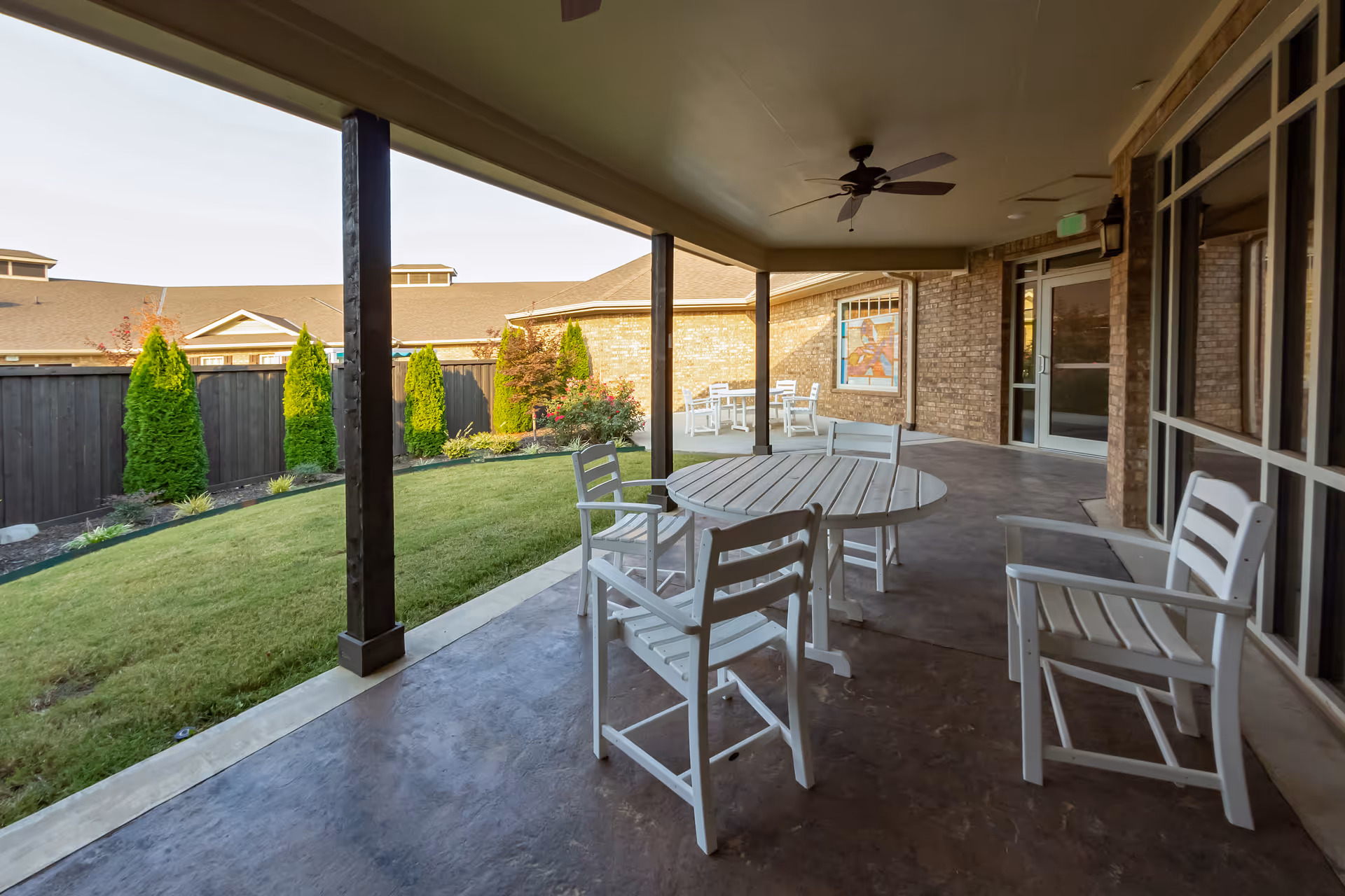Covered outdoor patio with white wooden chairs and round tables overlooking a small lawn and landscaped fenced yard.