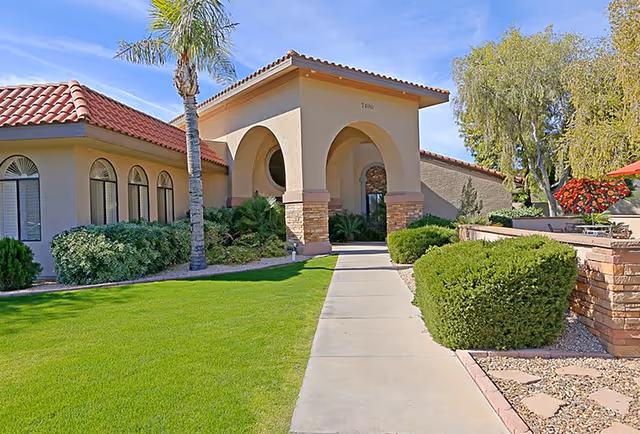 Exterior view of a senior living facility with a beige stucco building featuring arched entryways and a red tile roof. There is a concrete walkway leading to the entrance, surrounded by well-maintained green grass, bushes, palm trees, and other landscaping. The sky is clear and blue.