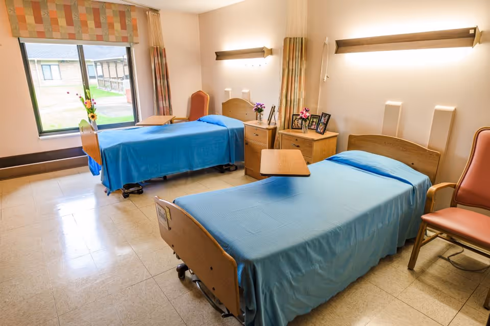 A bright room with two hospital-style beds covered in blue blankets, each with a wooden headboard and a small overbed table. There are two wooden nightstands between the beds, decorated with flowers and framed photos. A large window with patterned curtains lets in natural light, and there are two cushioned chairs near the beds. The floor is tiled and the walls are light-colored with mounted lights above each bed.