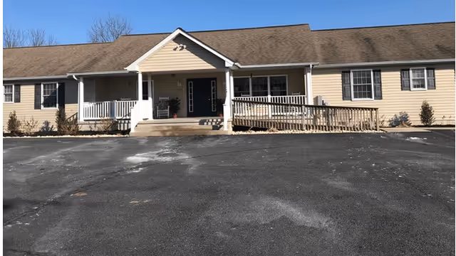 Single-story beige building with a brown shingled roof, a front porch with white railings, a wheelchair ramp, and a paved parking area in front under a clear blue sky.
