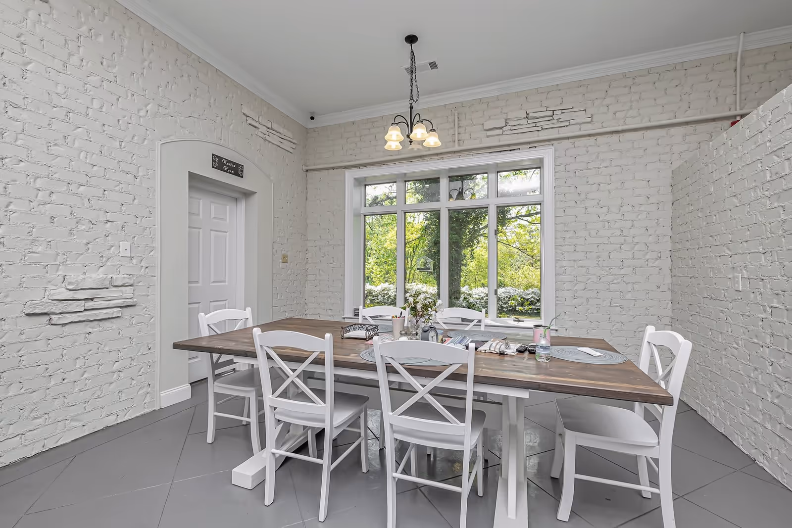 A bright dining room with white painted brick walls and a large window showing green trees outside. The room features a wooden dining table with six white chairs around it. On the table are placemats, a small vase with flowers, and some miscellaneous items. A chandelier with five lights hangs from the ceiling.