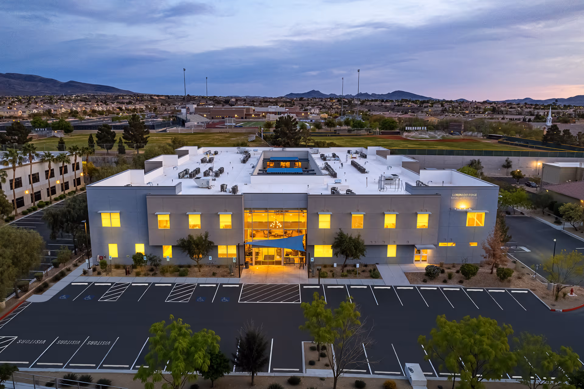 Aerial dusk view of the two-story Coronado Ridge Skilled Nursing and Rehabilitation Center with illuminated windows and an empty parking lot in front.