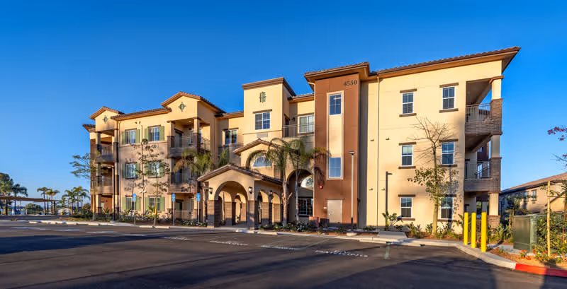 Exterior view of a three-story senior living facility building with a Mediterranean architectural style, featuring beige and brown walls, arched entryways, balconies, palm trees, and a parking lot in front under a clear blue sky.
