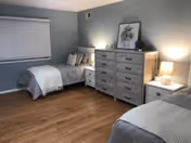 A cozy bedroom with two single beds, each with gray bedding and pillows. Between the beds is a white nightstand with a lamp. Against the wall is a wooden dresser with six drawers, decorated with a framed floral picture and a small clock. The room has light blue walls, a window with closed blinds, and wooden flooring.
