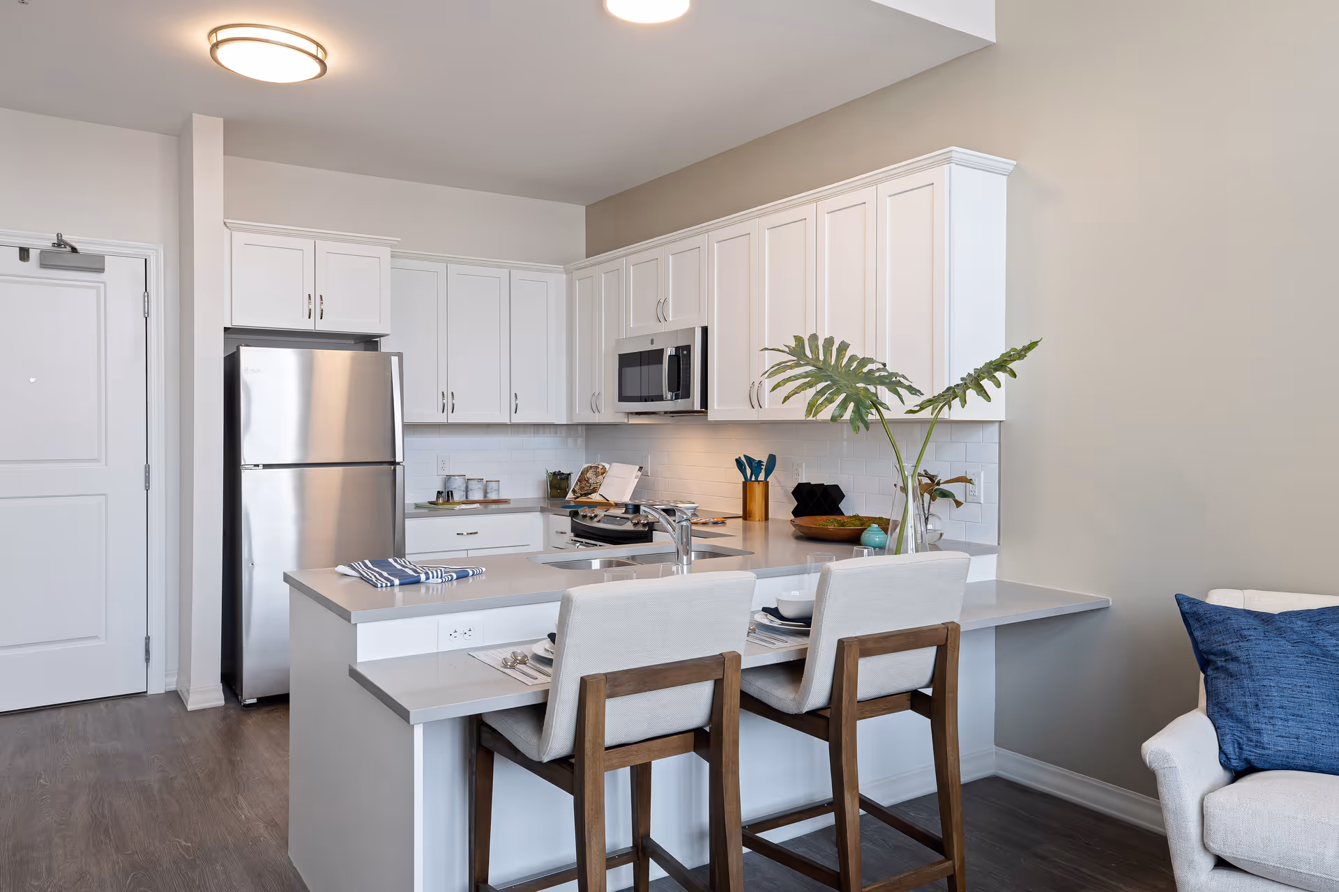 Modern kitchen area with white cabinets, stainless steel refrigerator and microwave, a countertop with a sink, two cushioned bar stools, and decorative plants. Adjacent to the kitchen is part of a living room with a white armchair and a blue pillow.