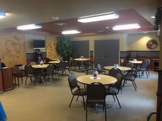 A communal dining area with several round tables, each surrounded by chairs. The room has beige walls with decorative artwork, a large potted plant, a wall-mounted TV, and overhead fluorescent lighting. There are double doors and additional seating along the walls.