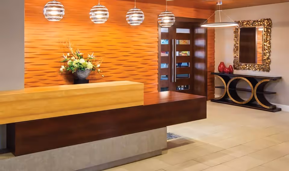 Modern reception area with a wooden front desk, three hanging spherical pendant lights, a textured orange wall with a floral arrangement, and a decorative console table with a large ornate mirror on the right side.