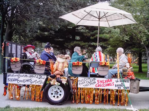 A group of elderly people sitting on a decorated trailer outdoors with trees in the background. The trailer is adorned with fall-themed decorations such as orange garlands and signs that read welcoming messages for Sylvan Crossings. A large white umbrella provides shade over the group.