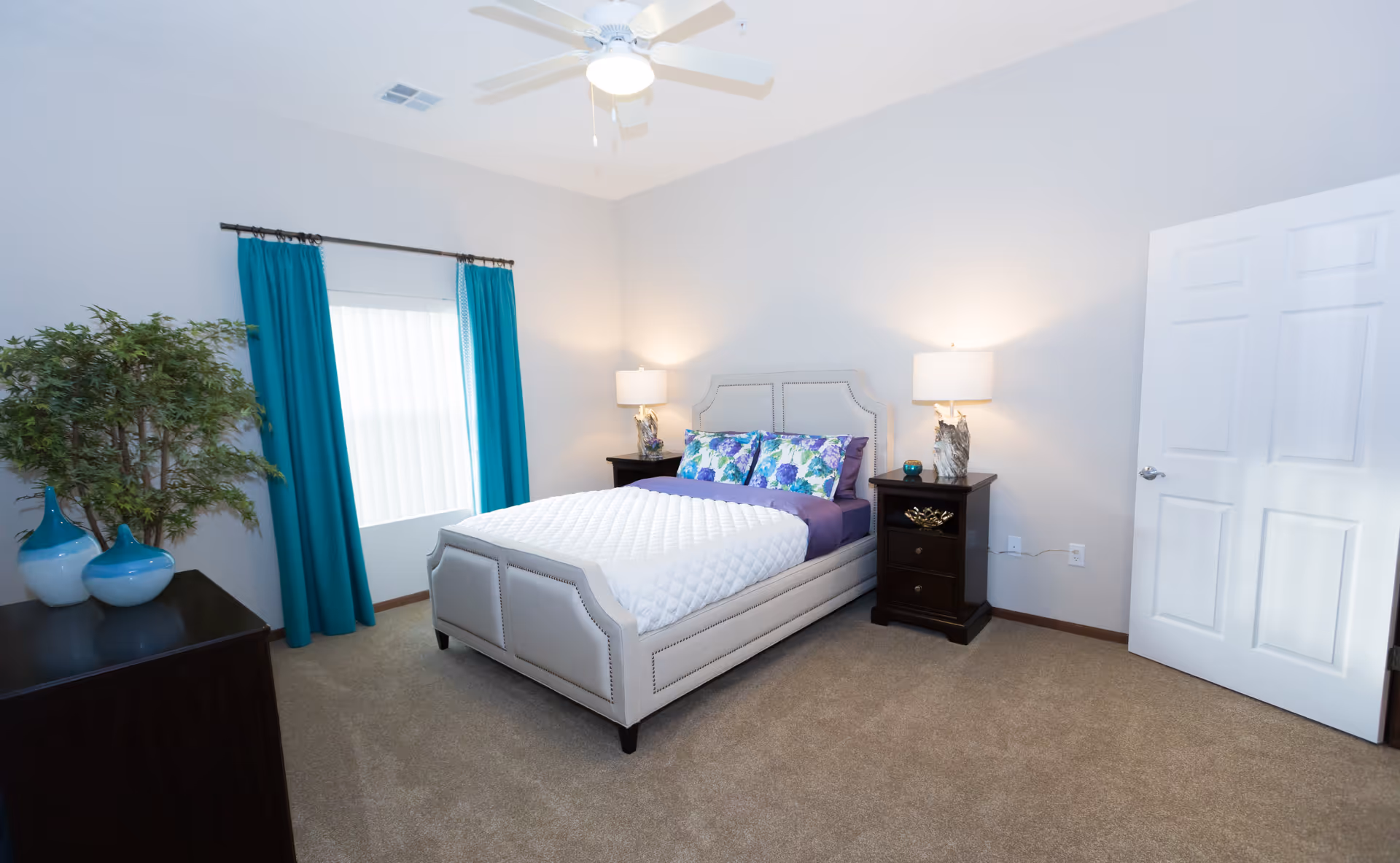 A neatly arranged bedroom with a bed featuring a white quilt and floral pillows, two dark wooden nightstands with lamps on either side, a window with teal curtains, a ceiling fan with light, a dresser with decorative vases, and a potted plant in the corner.