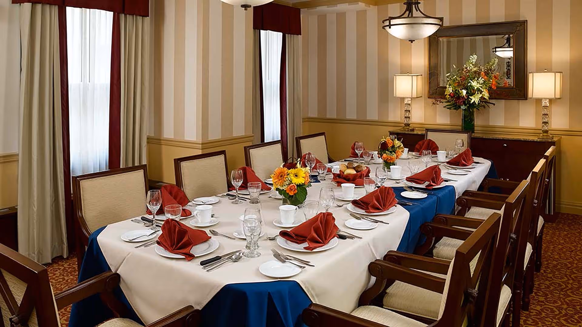 A formal dining room with a long table set for a meal. The table is covered with a white tablecloth and a blue runner, and is set with white plates, cups, glasses, silverware, and red folded napkins. There are flower arrangements placed along the center of the table. The room has beige and cream striped wallpaper, two windows with cream curtains and red valances, a large mirror on the wall, two table lamps, and a chandelier hanging from the ceiling.