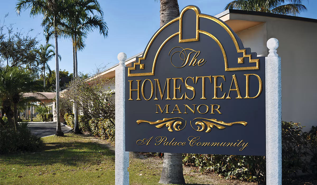 A large decorative sign for The Homestead Manor, a palace community, standing outdoors with palm trees and bushes in the background.