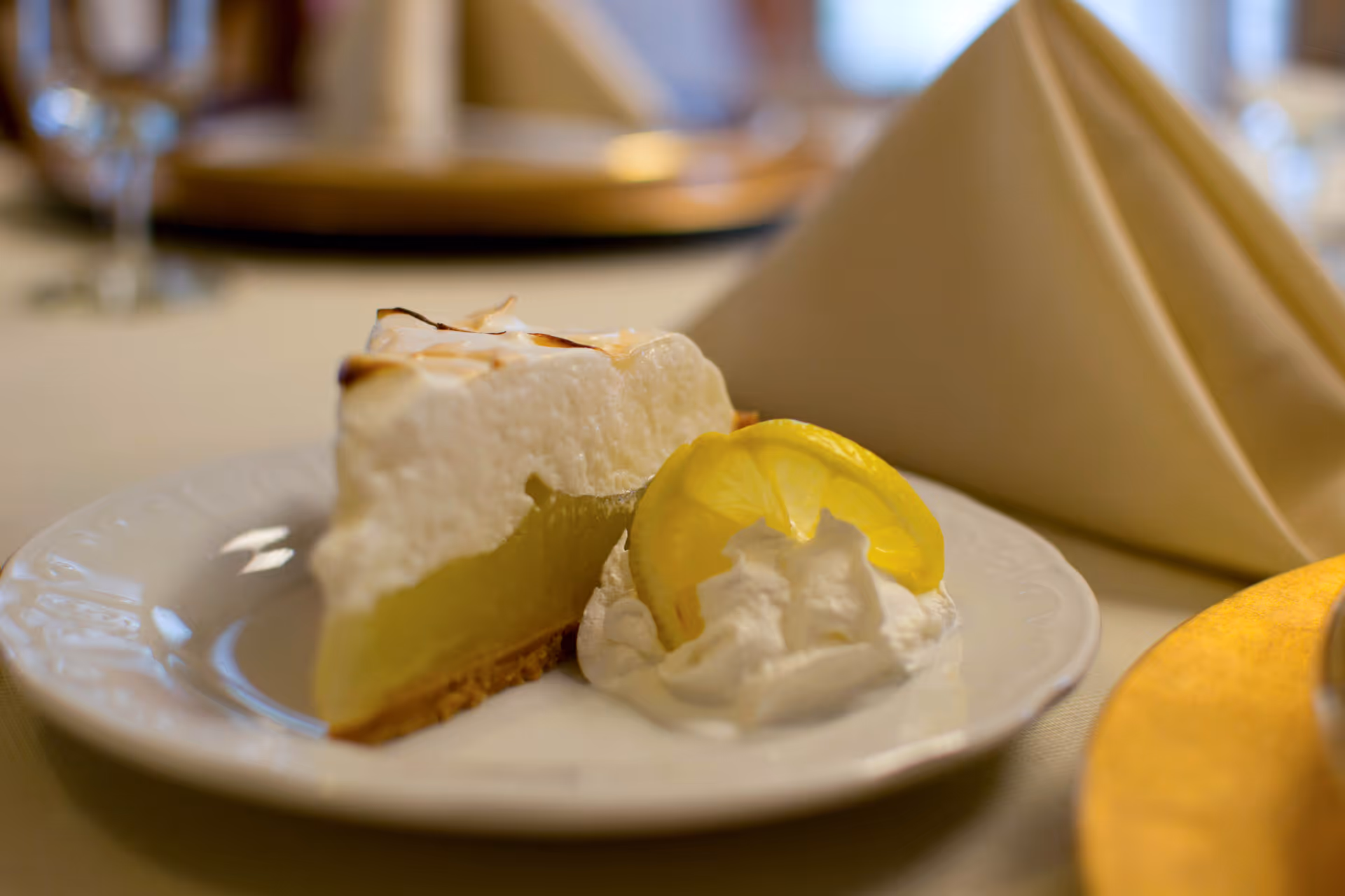A slice of lemon meringue pie with whipped cream and a lemon wedge on a white plate at a table setting.