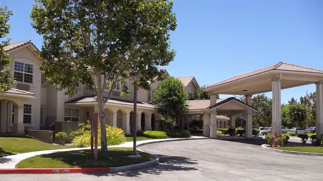 Front entrance of a senior living building with a covered porte-cochere, driveway, landscaping, and trees under a clear sky.