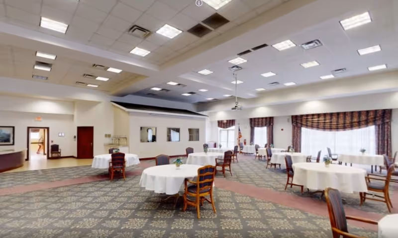 Spacious dining/activity room with round tables covered in white tablecloths, wooden chairs, patterned carpet, and large windows with drapes.