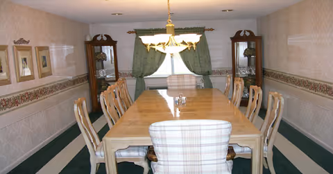 A formal dining room with a long wooden table surrounded by ten chairs, two of which have plaid upholstery. The room features patterned wallpaper with a floral border, green curtains framing a window at the far end, two glass-fronted display cabinets, and a chandelier hanging above the table.