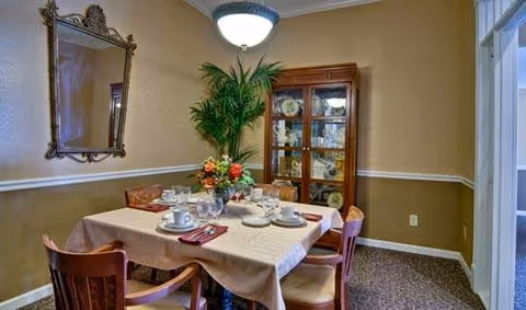 A cozy dining room with a table set for four, featuring white plates, cups, and silverware on a beige tablecloth. There are four wooden chairs around the table. A tall green plant is placed in the corner next to a wooden china cabinet displaying plates and glassware. A decorative mirror hangs on the wall above the plant, and a ceiling light fixture illuminates the room.