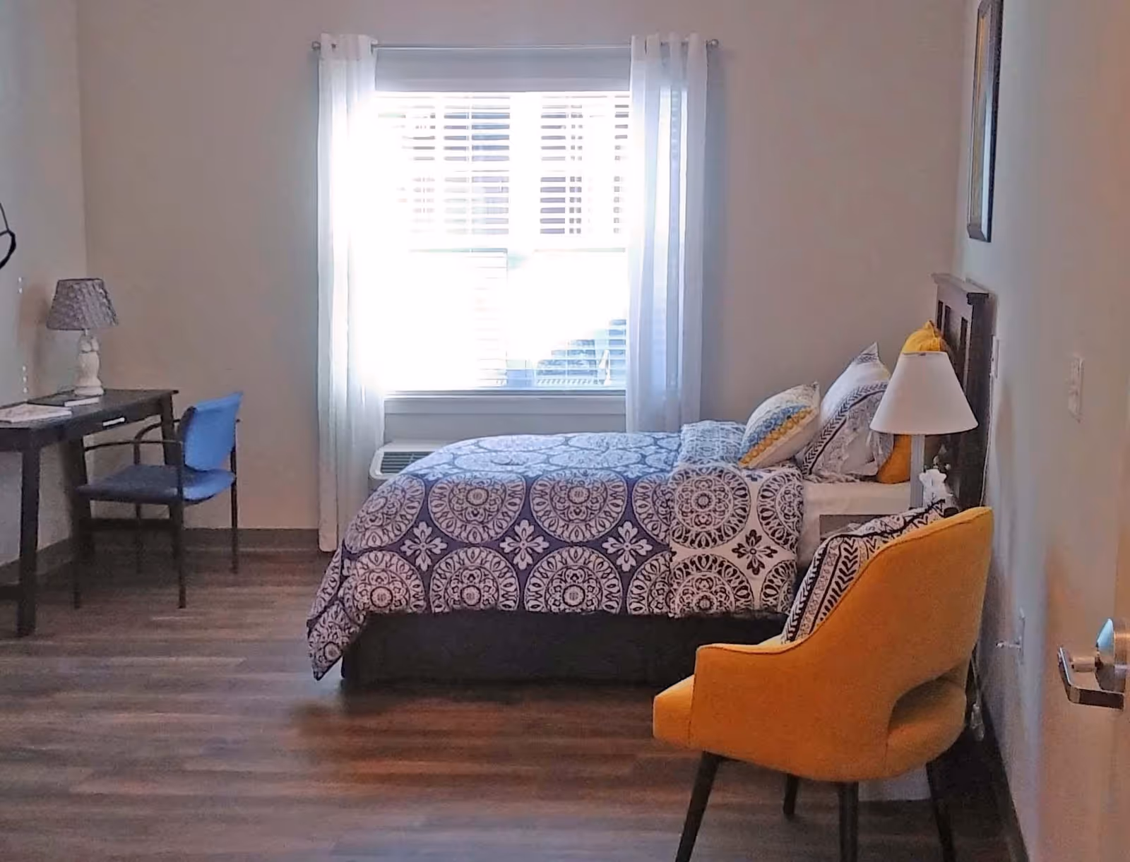 A bedroom with a bed covered in a blue and white patterned comforter and pillows, a yellow armchair with a patterned cushion, a small white nightstand with a lamp, a desk with a chair, and a window with white curtains letting in natural light.
