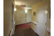 Interior hallway of a retirement home with patterned carpet, white doors, handrails, and wall lighting.