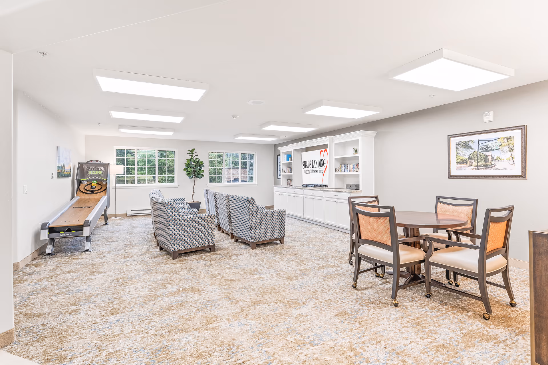A bright and spacious common area in a senior living facility featuring a row of patterned armchairs facing a large built-in white entertainment center with a TV displaying the Shads Landing Gracious Retirement Living logo. To the right, there is a round wooden table with four chairs. On the left side, there is a skee-ball game machine near two large windows letting in natural light. The room has beige carpet and white walls with framed artwork and a tall potted plant.