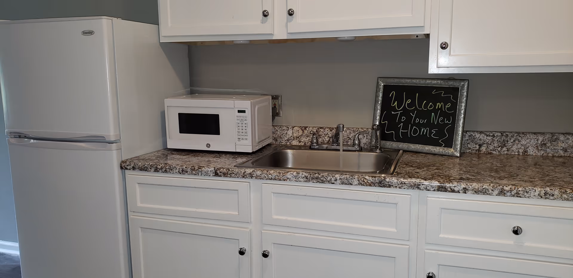 A kitchen area with white cabinets, a white refrigerator, a white microwave on a granite countertop, and a stainless steel sink with running water. A small framed chalkboard on the counter reads 'Welcome To Your New Home'.
