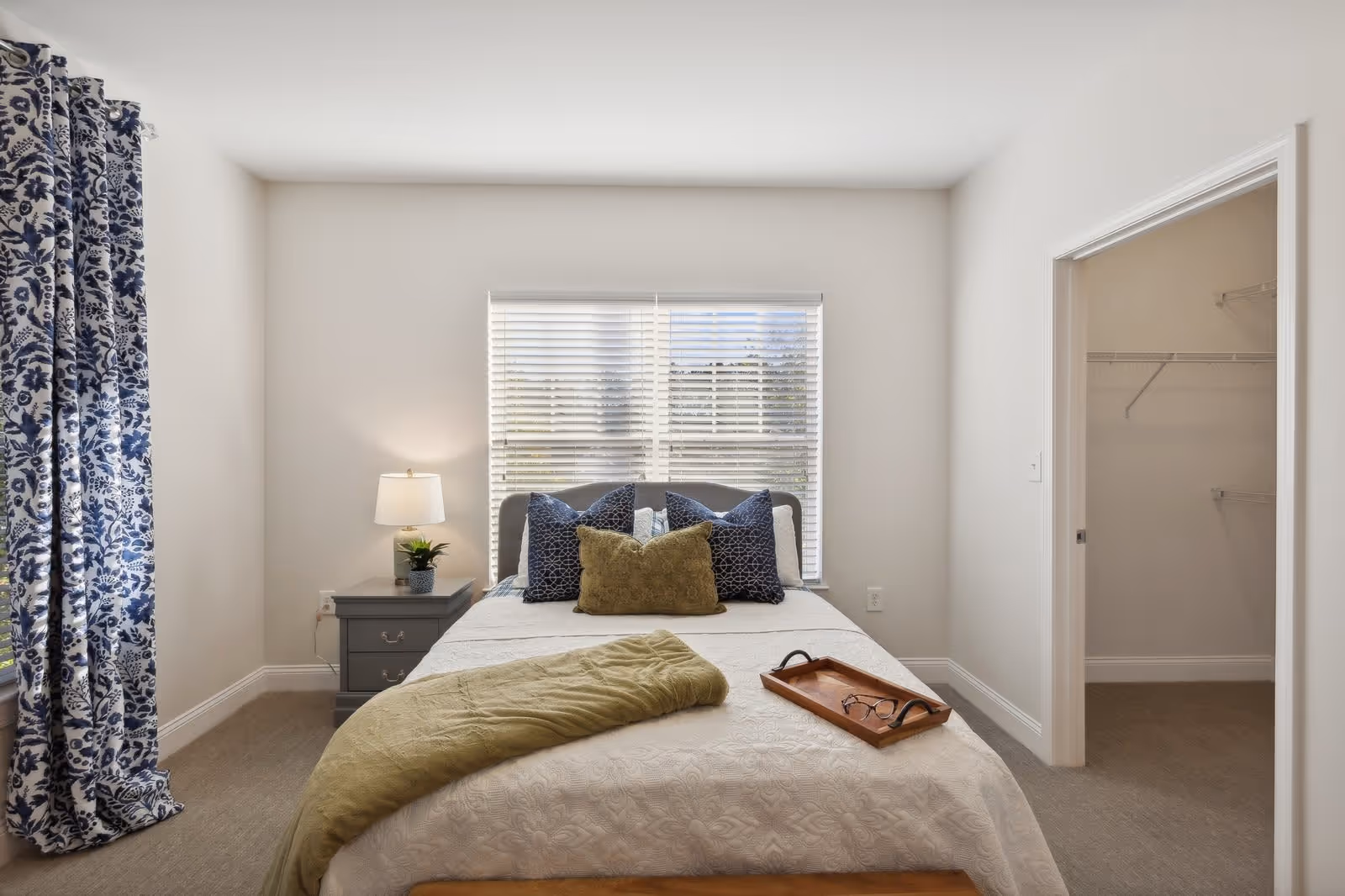 A bright bedroom with a neatly made bed featuring white bedding, green and blue pillows, and a green throw blanket. There is a wooden tray with glasses on the bed. To the left of the bed is a gray nightstand with a lamp and a small plant. A window with white blinds is centered behind the bed, and blue and white patterned curtains hang on the left side. To the right, there is an open doorway leading to a walk-in closet with wire shelving.