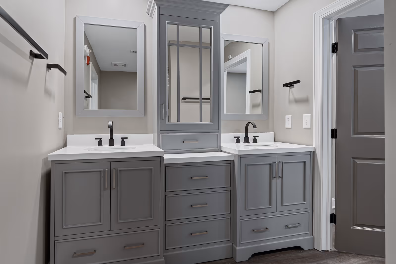 A modern bathroom vanity with two sinks, each with black faucets and soap dispensers. The vanity is painted gray with multiple drawers and cabinets, and there are two rectangular mirrors mounted above each sink. A tall cabinet with glass-paneled doors is centered between the sinks. The walls are painted light gray, and there are black towel bars on either side of the vanity. A gray door is visible on the right side of the image.